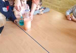 Na stole stoi szklanka z pomarańczową wodą, soda, mleko z pomarańczowym barwnikiem. Pani nabiera w strzykawkę niebieska wodą. Dzieci siedzą wokół stołu.