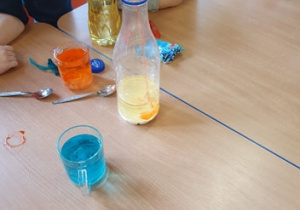 Na stole stoi szklanka z wodą zabarwioną na niebiesko, szklanka z wodą zabarwioną na pomarańczowo, olej, łyżeczki małe. Pani nalewa pomarańczową wodę do mleka. Chłopiec obserwuje nalewanie.