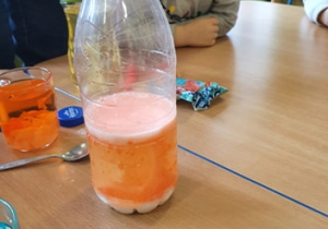 Na stole stoi woda zabarwiona na pomarańczowo, butelka z mlekiem zabarwionym na pomarańczowo. Chłopiec obserwuje butelkę.