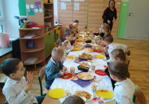 Dzieci siedzą przy stole, przykrytym białym obrusem. Na nim znajdują się słodkości i soki. Na końcu stołu stoi pani.