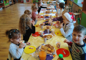 Dzieci siedzą przy stole, przykrytym białym obrusem. Na nim znajdują się słodkości i soki.