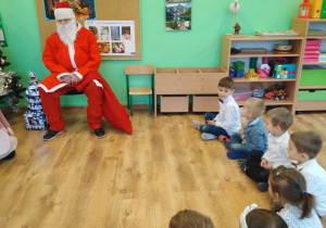 Pod ścianą na krześle siedzi święty Mikołaj. Na podłodze siedzą dzieci w kole.