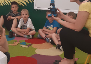 Z lewej strony siedzi chłopiec, pod ścianą siedzi 3 chłopców oraz Pani nauczycielka, która trzyma w ręce mikroskop