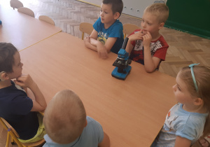 Przy stole od lewej strony siedzi 2 chłopców, dziewczynka, chłopiec, na stole stoją 2 mikroskopy