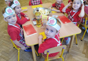 Dzieci w czerwonych koszulkach i fartuszkach siedzą przy stole z produktami do pieczenia