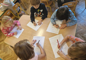 Dzieci siedzą przy stole i kolorują postać listonosza