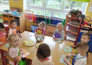 Dzieci siedzą przy stole i kroją owoce.