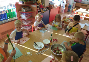 Dzieci siedzą przy stole i kroją składniki na sałatkę.