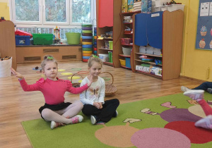Dzieci siedzą na dywanie z woreczkami gimnastycznymi umieszczonymi na głowie.