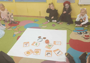 Dzieci siedzą na dywanie. Na środku jest plansza z flascards.