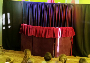 Dzieci siedzą na widowni i oglądają przedstawienie teatralne. N scenie widać kukiełkę czarownicy