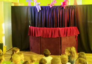Dzieci oglądają przedstawienie teatralne. Na scenie widać 3 kukiełki
