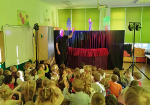 Dzieci oglądają przedstawienie teatralne. Po lewej stronie stoi aktor, po prawej stronie widać kukiełkę przedstawiającą chłopca.