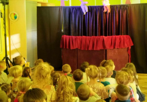 Dzieci siedzą na widowni i oglądają przedstawienie teatralne, w tle widać 2 kukiełki