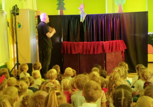 Na zdjęciu widać dzieci oglądające przedstawienie teatralne. Przed nimi stoi aktor ubrany na czarno.
