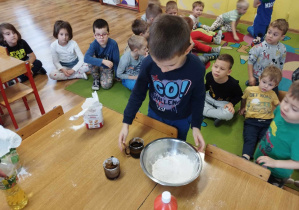 Chłopiec wlewa olej do miski. W tle dzieci siedzą na dywanie.