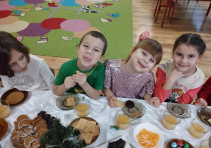 Dzieci siedzą przy stole i częstują się owocami i słodyczami.