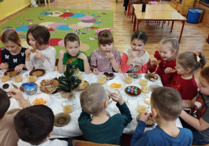 Dzieci siedzą przy stole i częstują się owocami i słodyczami.