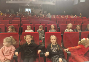 Dzieci siedzą na czerwonych fotelach w oczekiwaniu na sztuke teatralną