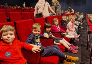Dzieci siedzą na czerwonych fotelach w oczekiwaniu na sztuke teatralną
