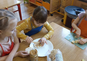 Dzieci siedzą przy stole, smarują wafle ryżowe masłem orzechowym i kroją banana.