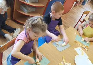 Dzieci siedzą przy stole i kroją banany.
