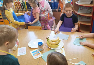 Dzieci stoją wokół stołu, na którym są składniki do zrobienia wafli ryżowych z masłem orzechowym i bananem.