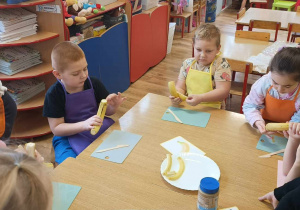 Dzieci siedzą przy stole i obierają banany.