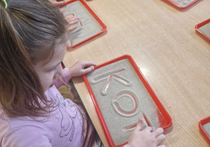Dzieci piszą na tackach z piaskiem wyraz kot