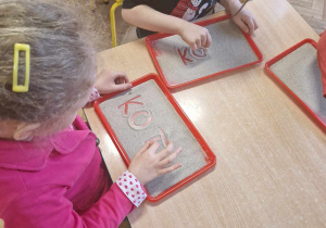 Dzieci piszą na tackach z piaskiem wyraz kot