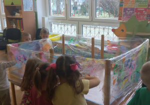 Dzieci stoją przy stole, który jest owinięty folią i malują farbami za pomocą paluszków.