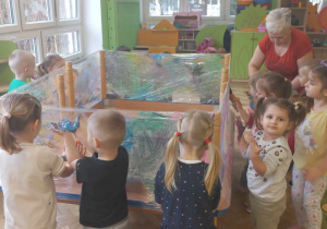 Dzieci stoją przy stole, który jest owinięty folią i malują farbami za pomocą paluszków.