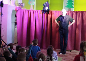 Dzieci oglądają przedstawienie teatralne, na scenie widać aktora w czarnej koszuli oraz kukiełkę kota